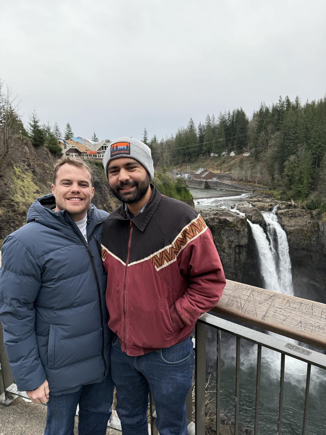 Two men wearing coats standing on a scenic overlook, facing the camera and smiling. Behind them, a large waterfall and a lodge structure, made famous by the Twin Peaks intro, are visible.