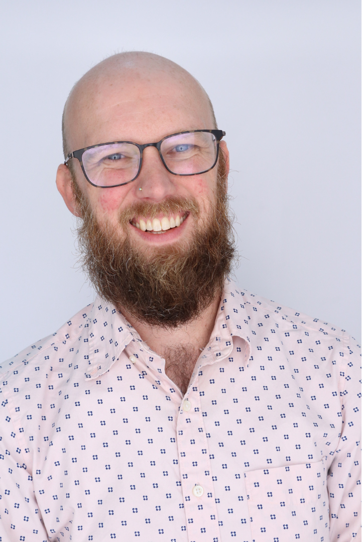 A headshot of Elliot Williams, a smiling bald white man with a beard and glasses, against a neutral grey background.