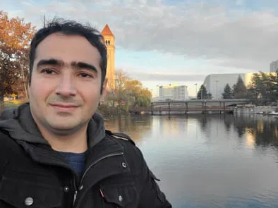 A selfie of Seyed Abdollah Shahrokni. He is standing outside in a black jacket at Riverfront Park in Spokane, WA. Behind him is the Spokane River and the iconic Great Northern Clocktower on an overcast day.
