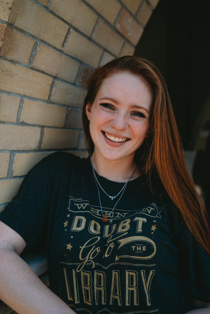 A photo of Isabelle Antes in a grey shirt that reads: "When in doubt, go to the library" standing in a brick archway.