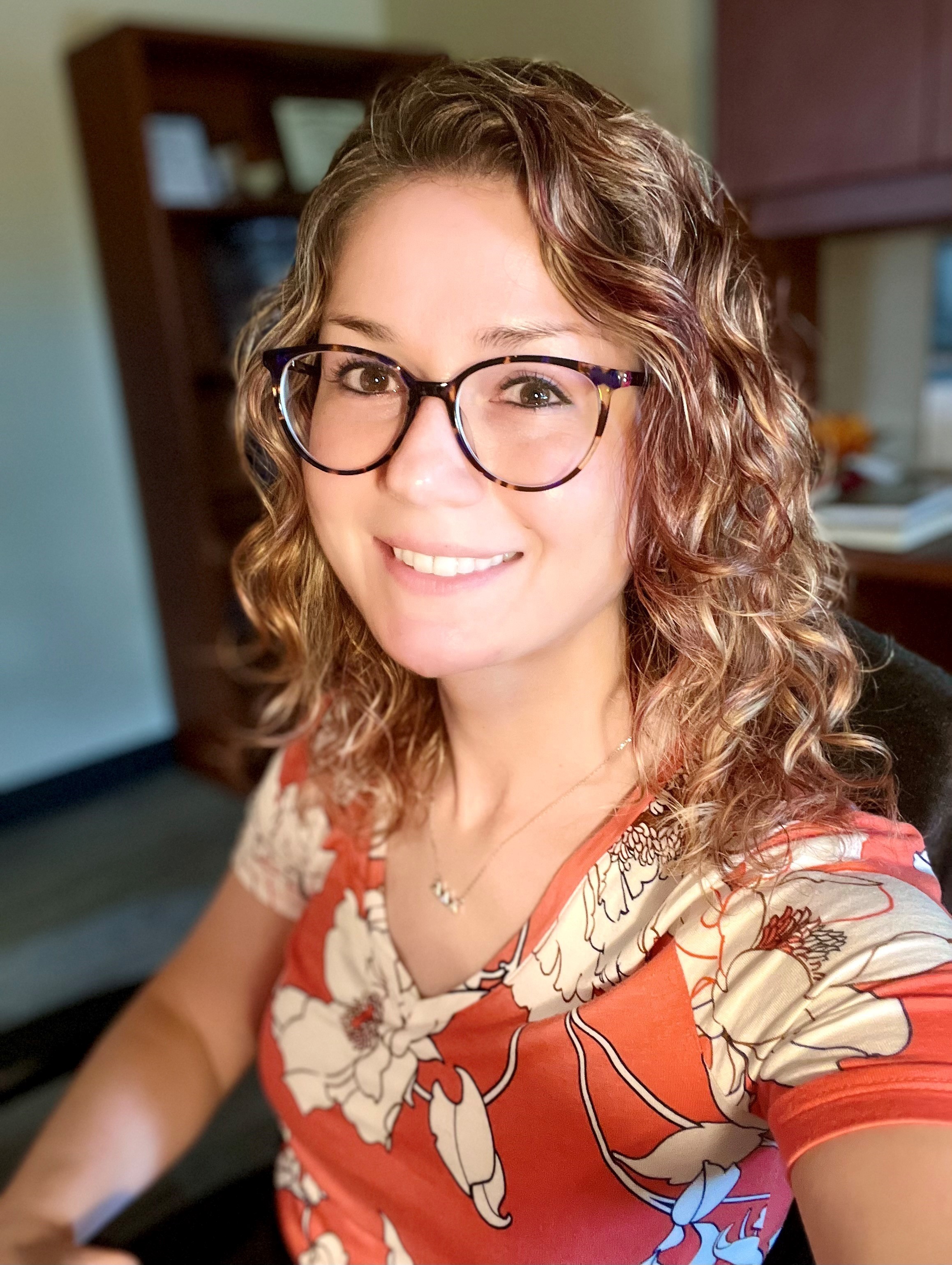 A headshot of Millicent Weber with curly reddish-brown hair in an orange shirt against a dark office background.