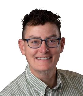 A headshot against a white background of a smiling person with a tannish-pink complexion and freckles. Their dark brown hair is close cropped on the sides with short open curls on top. Their light-colored eyes are surrounded by blue-rimmed eyeglasses, and their chin sprouts a few wispy gray hairs. Their button-down shirt has narrow vertical green and white stripes. They have no visible tattoos, piercings, or jewelry.